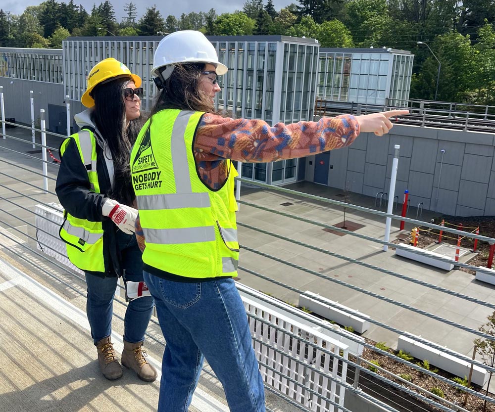 Two PRR staff in hard hats and high-visibility vests survey a rooftop construction site, one pointing across the building.