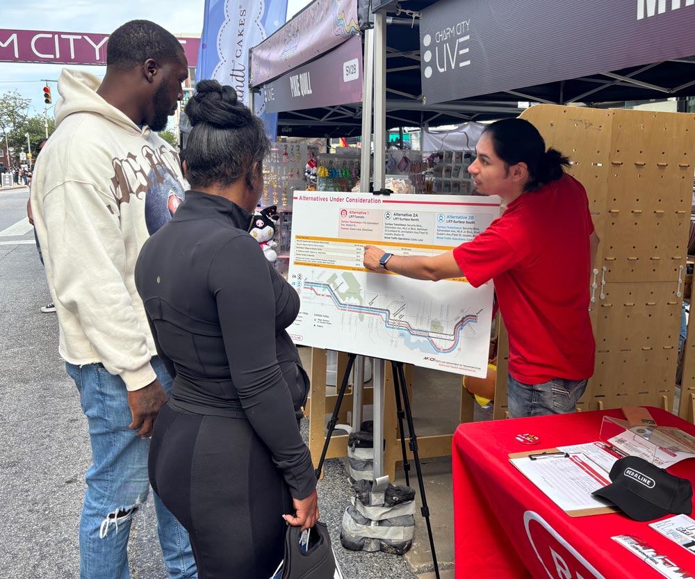 A PRR staff member walks two community members through a project alternatives map at a street outreach table.