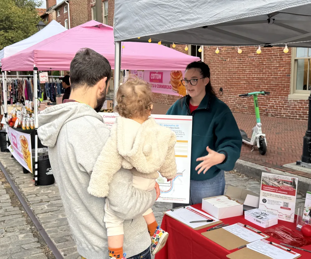 A PRR staff member talks with a parent holding a toddler at a Red Line outreach table at an outdoor street market.