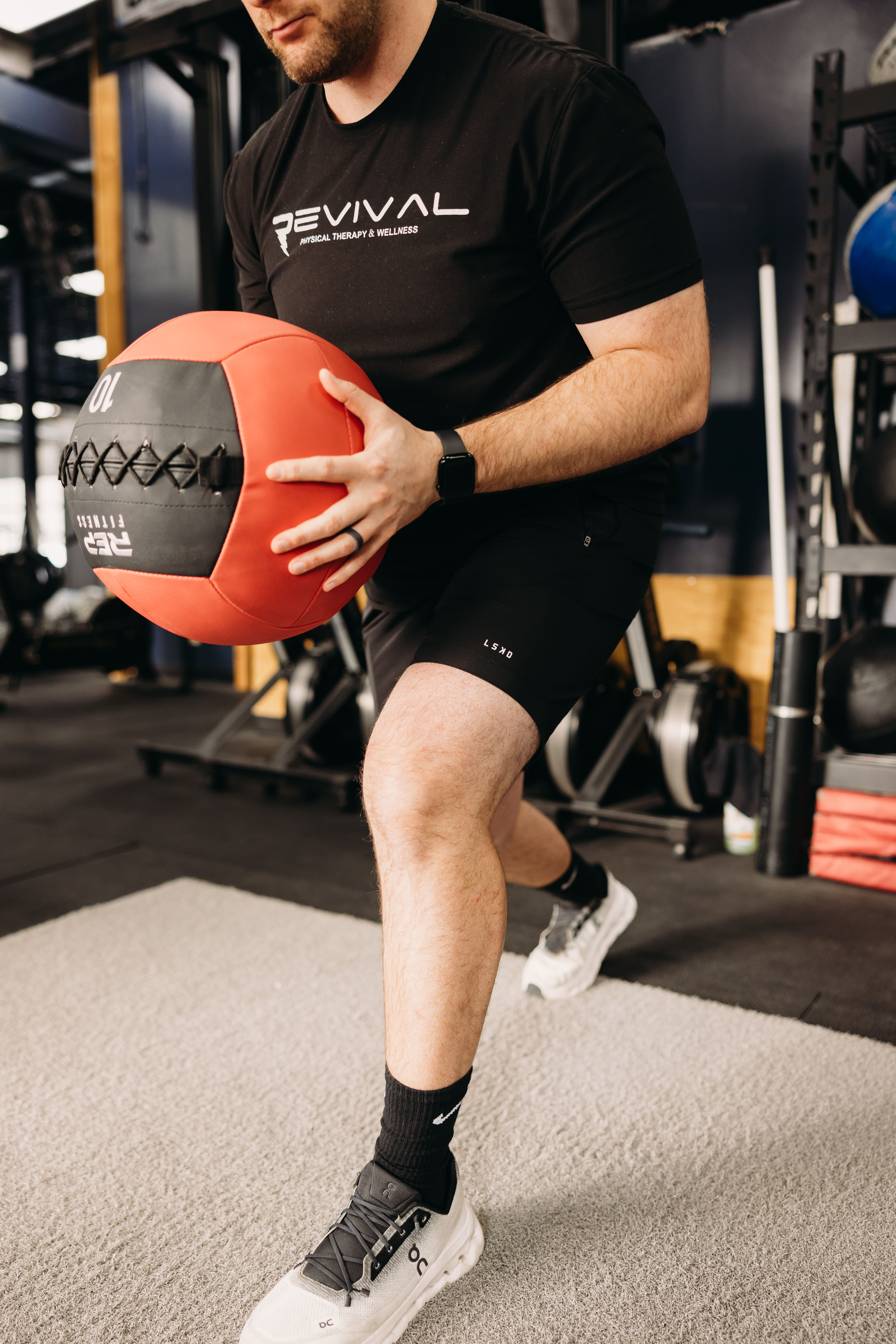 A trainer wearing a Revival Physical Therapy t-shirt performs a lunge in the gym while holding a red REP Fitness medicine ball