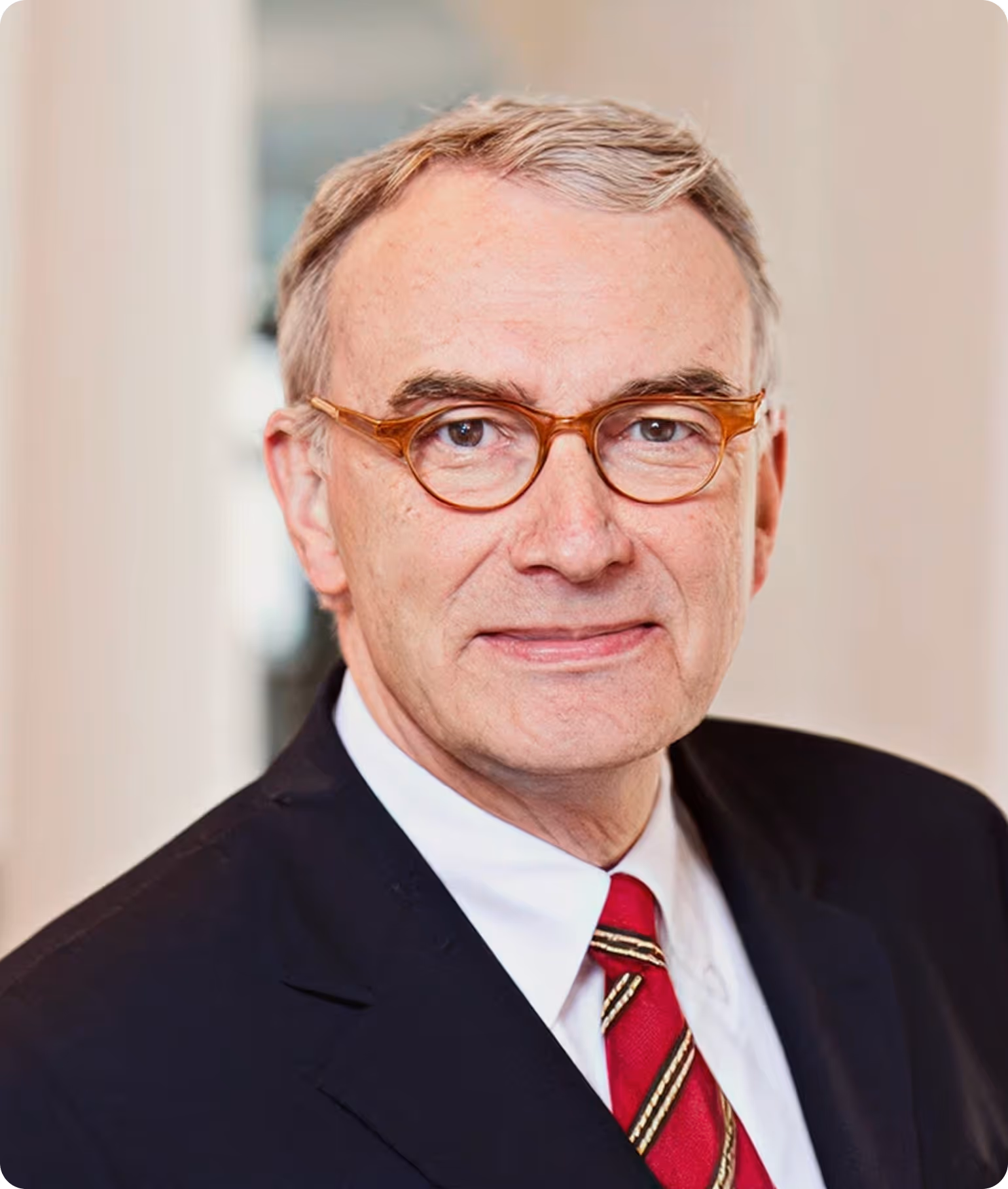 Älterer Mann mit hellbraunen Brillen, grauem Haar, Anzug und rot gestreifter Krawatte.