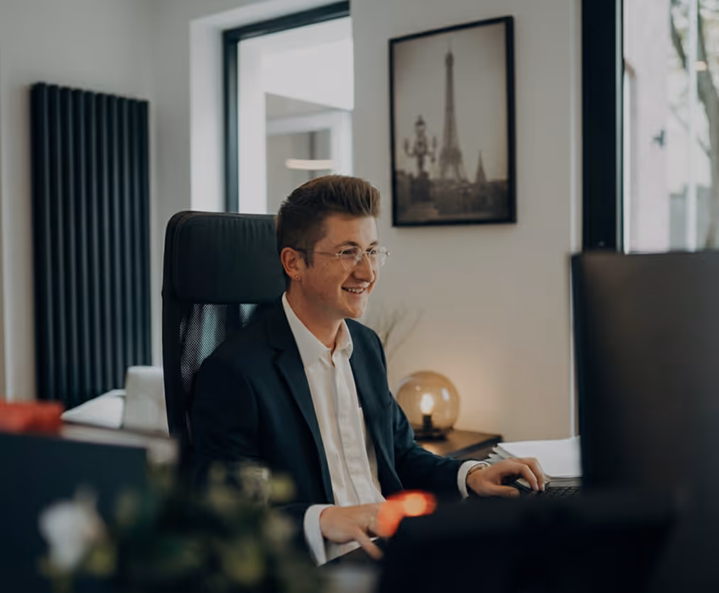 Junger Mann mit Brille sitzt lächelnd in einem Büro vor einem Computerbildschirm.