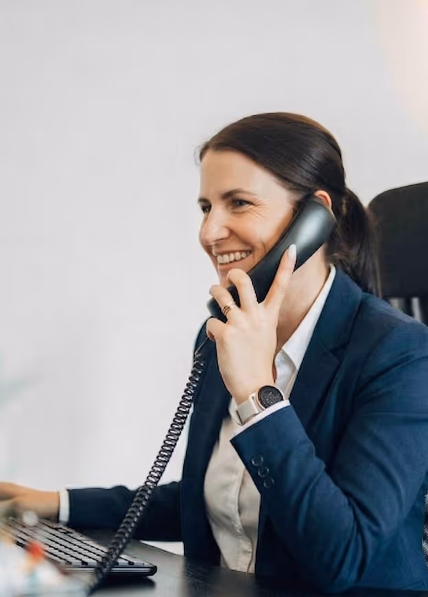 Frau in blauem Blazer spricht lächelnd am Schreibtisch in ein Telefon mit Schnur.