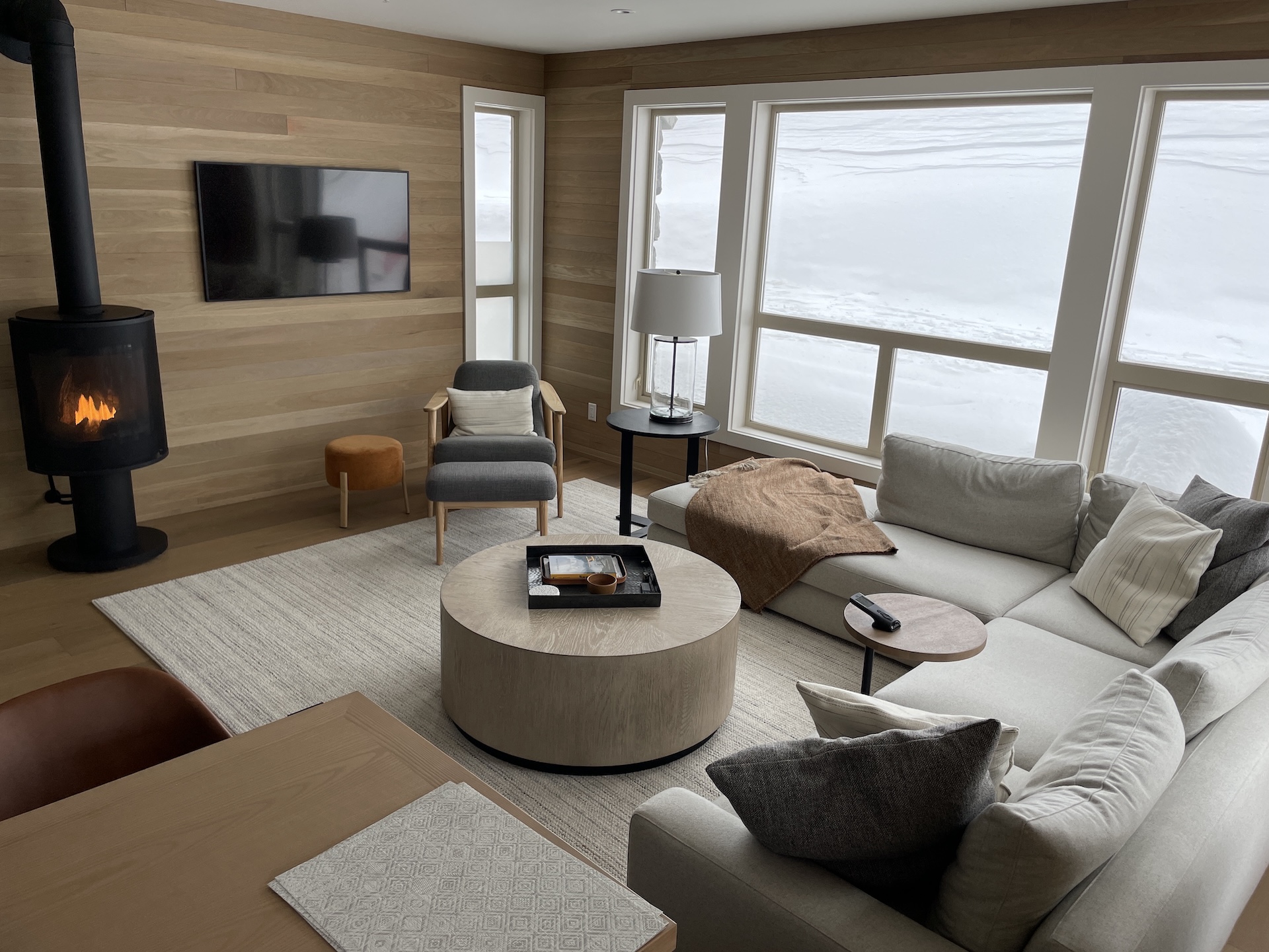 Cozy living room with wood walls, modern fireplace, gray sectional, armchair, and large snowy windows.