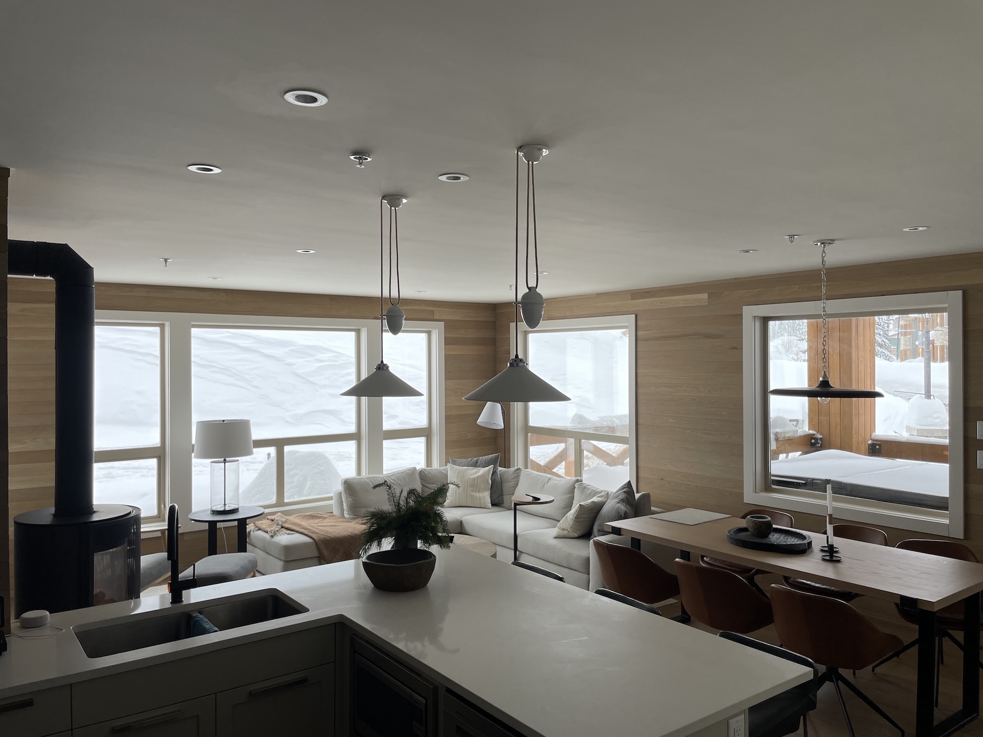 Cozy open-plan living area with kitchen island, white sectional sofa, wooden dining table, and snowy windows.