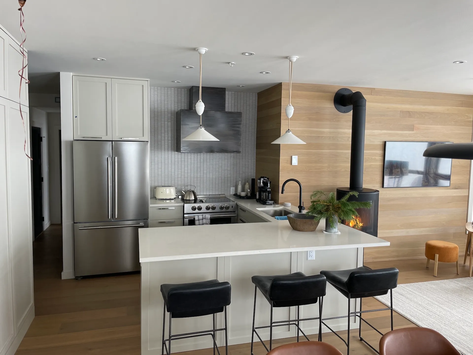 Modern kitchen with stainless steel fridge, white island, black stools, wood-paneled wall, and fireplace.