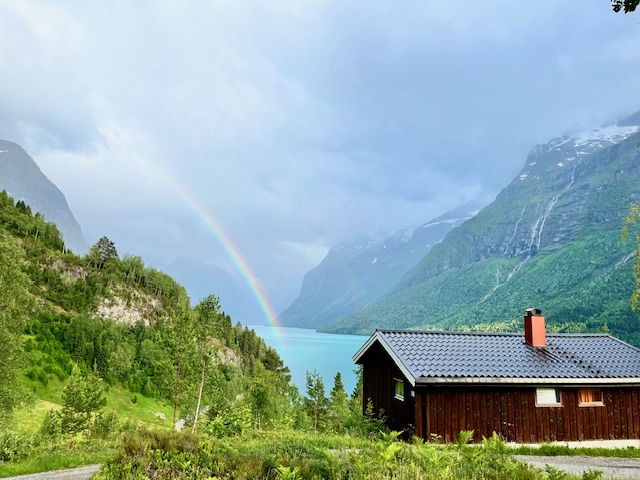 A wooden cabin sits in the foreground of a Norwegian fjord, with turquoise water and a rainbow in the background, framed by lush green mountains under a cloudy sky.