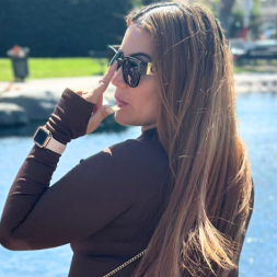Woman with long hair wearing sunglasses and dark top by poolside on a sunny day.