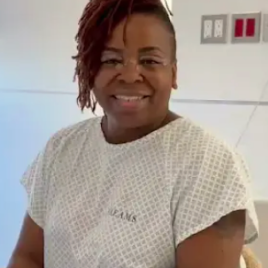 Smiling person wearing a hospital gown sitting indoors with a light background.