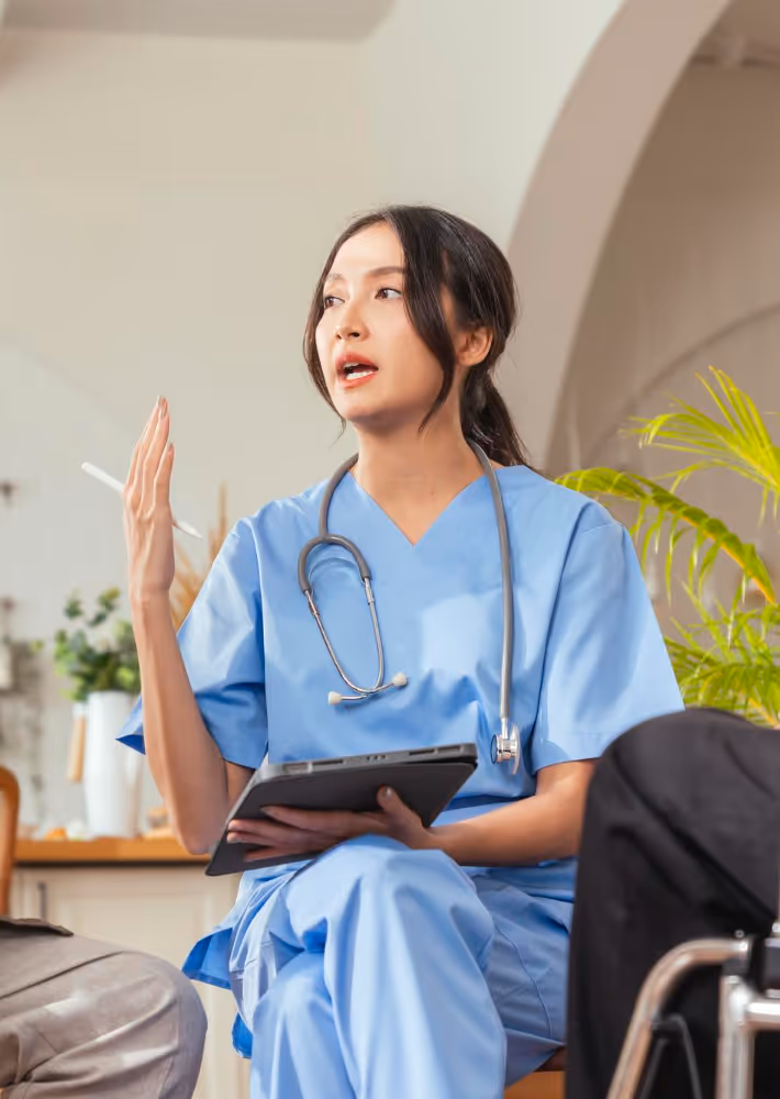 Female healthcare worker in blue scrubs holding tablet and gesturing while talking to seated people.