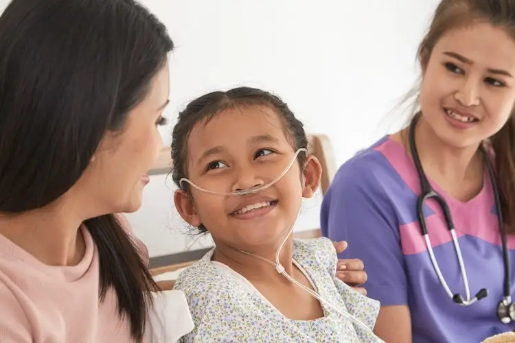 Smiling child with nasal oxygen tube looking at woman as nurse watches in purple scrubs with stethoscope.