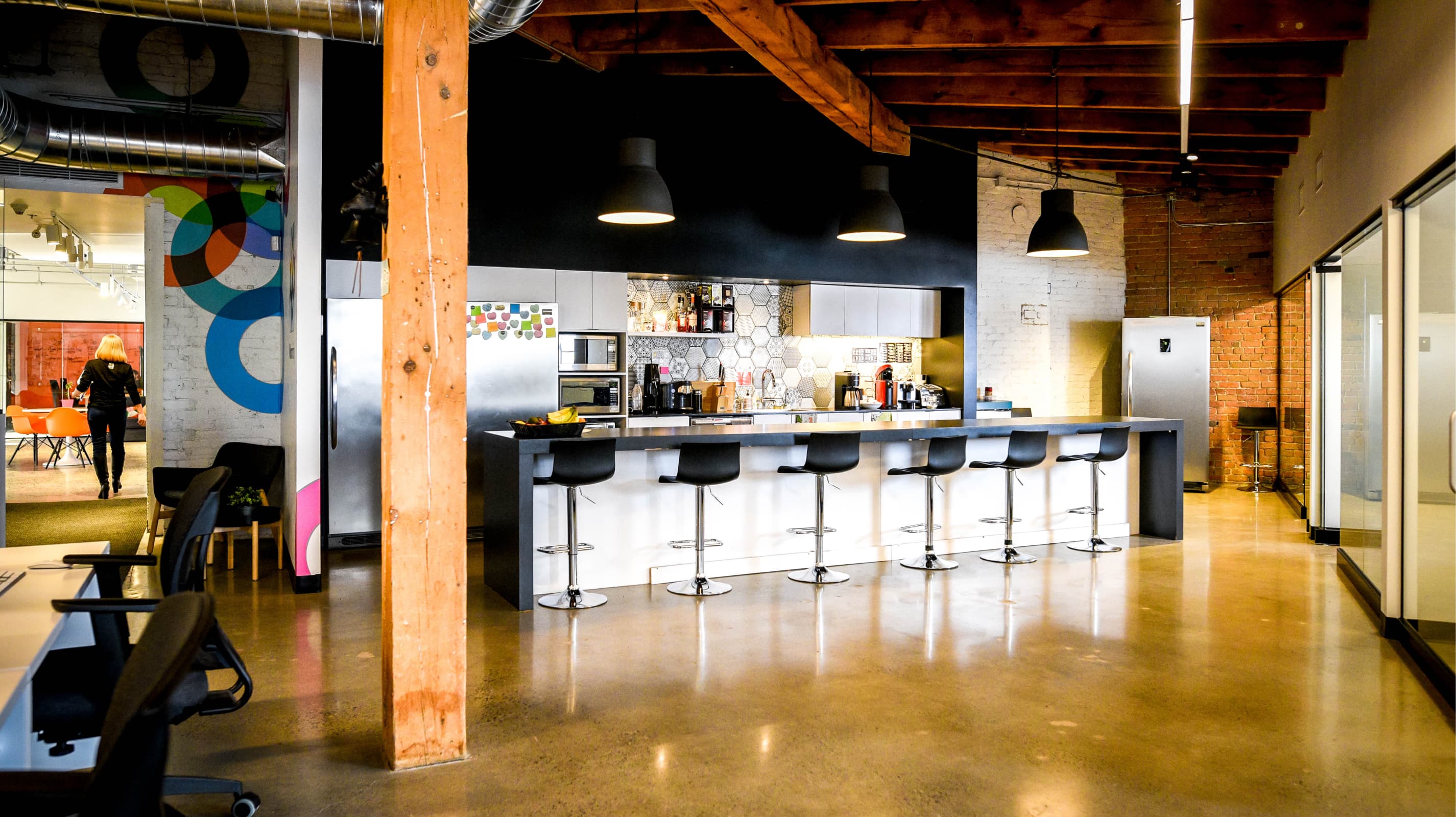 Modern office kitchen with black stools, wooden beams, brick walls, and a person walking in the background.