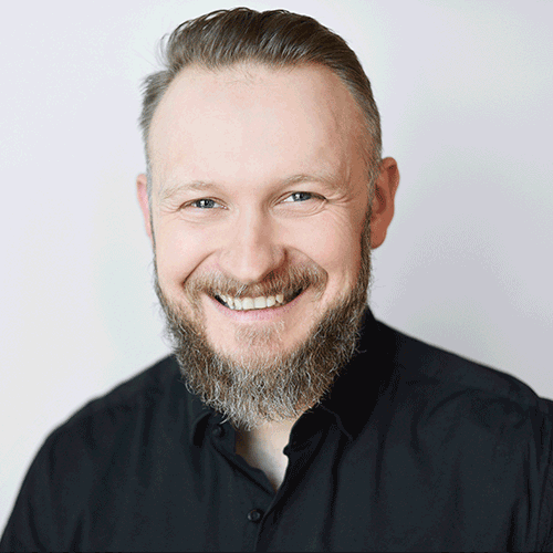 Worximity employee. Smiling man with beard and short hair wearing a black button-up shirt against a light background.