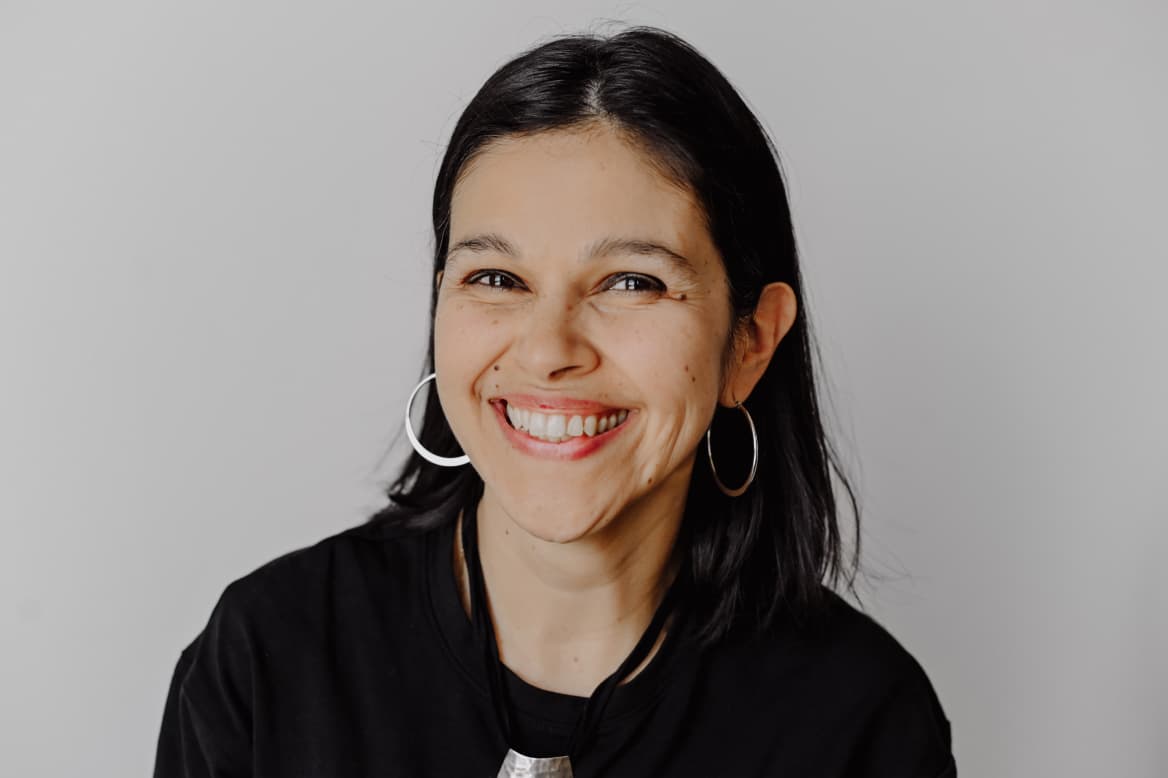 Worximity employee. Smiling woman with dark hair wearing hoop earrings and a black top against a plain background.