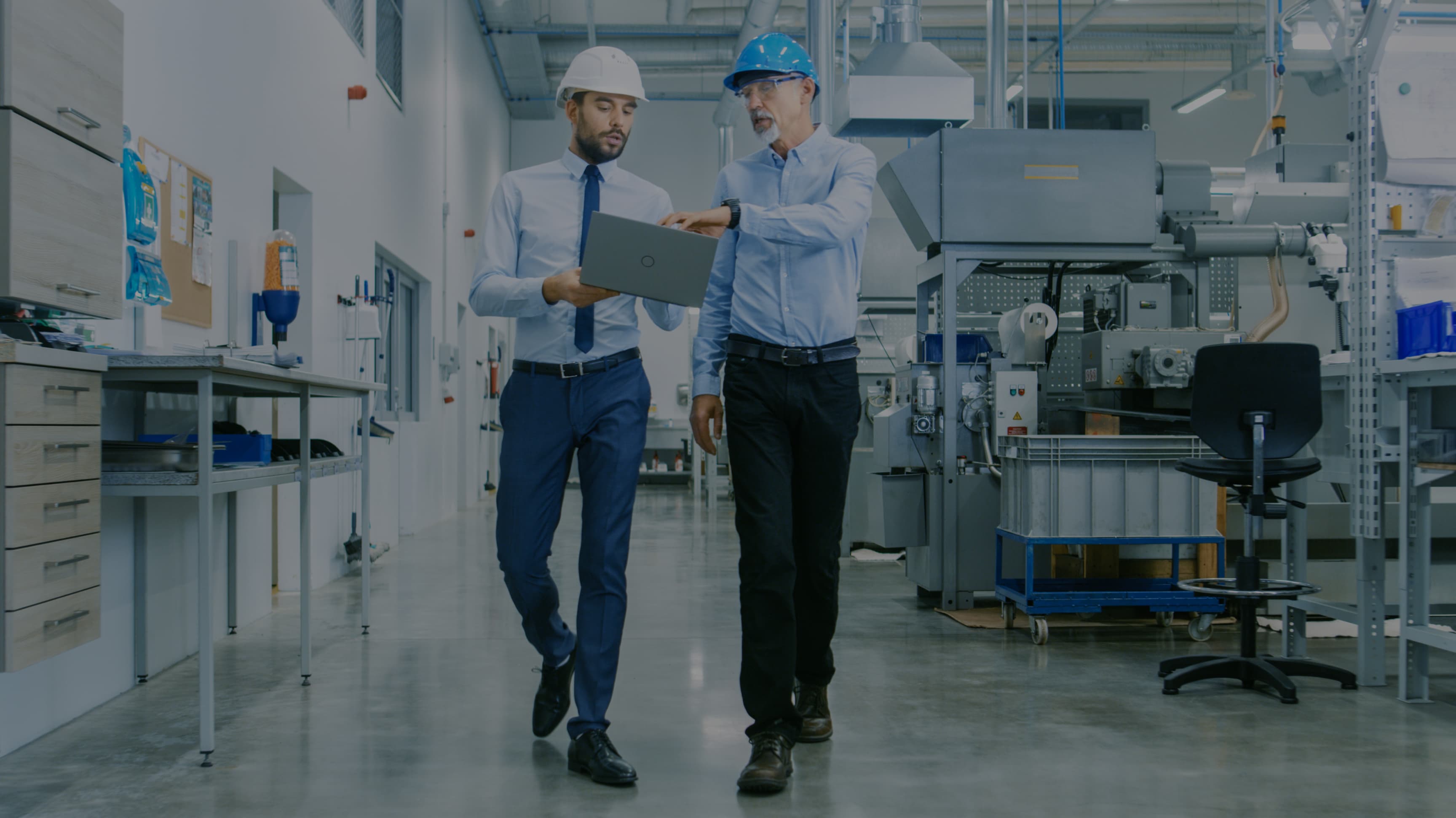 Two men in hard hats reviewing a laptop while walking in a manufacturing facility.