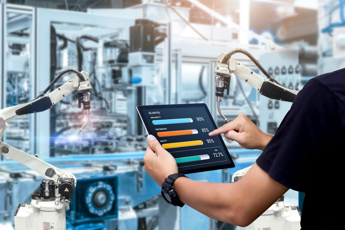 Person holding tablet showing manufacturing metrics with robotic arms in a factory background.