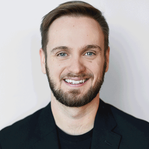 Smiling man with light brown hair and beard wearing a black jacket and shirt against a light background.