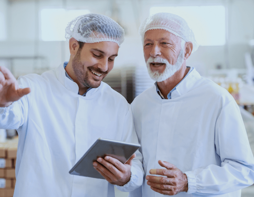 Two male workers in white coats and hairnets looking at a tablet and smiling.