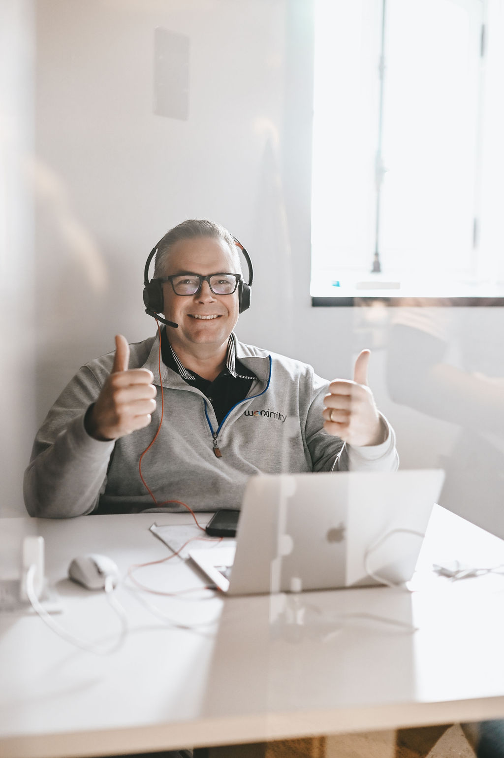 Smiling man with headset and glasses giving two thumbs up sitting at desk with laptop in bright room.