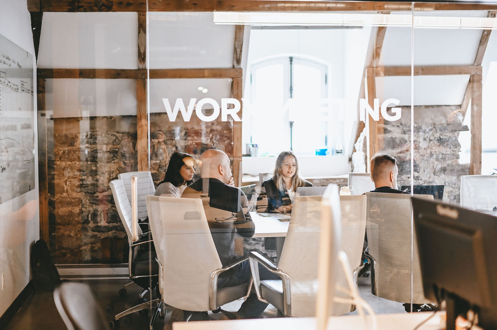Four people having a meeting inside a glass-walled conference room with exposed brick walls.