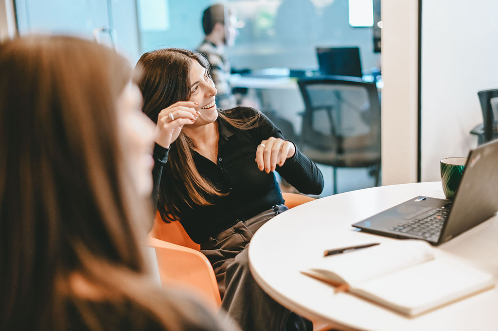 Smiling woman sitting at round table with laptop, notebook, and pen in a modern office setting.