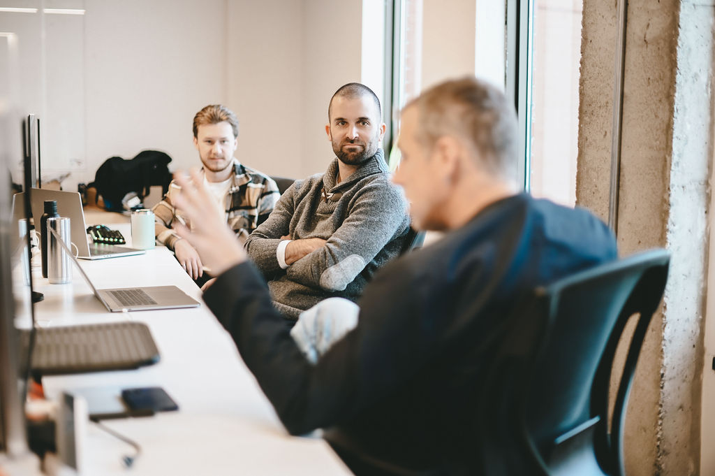 Three men in an office having a discussion, one in foreground gesturing while others listen.