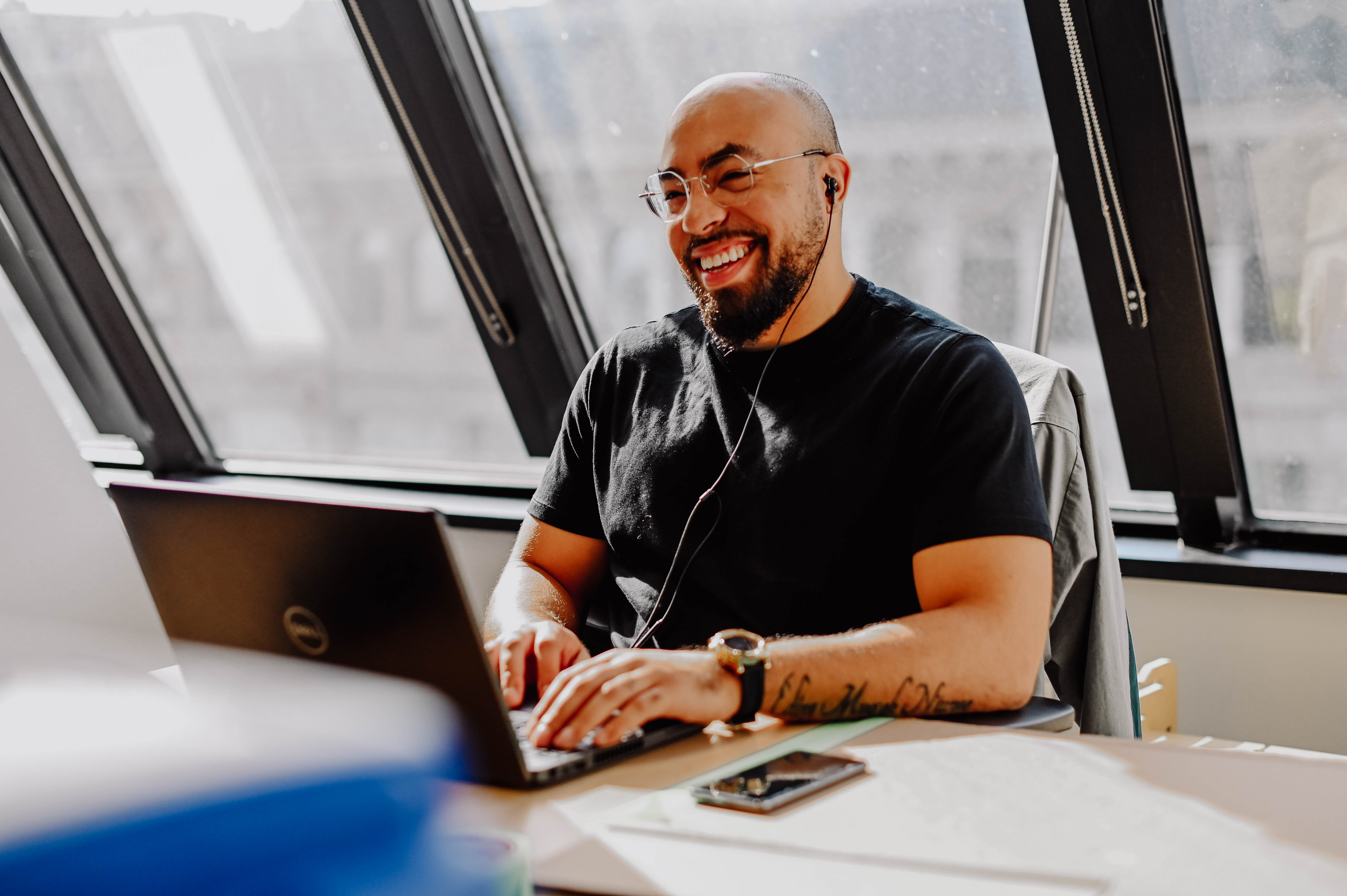 Smiling man with glasses and beard working on a laptop at a desk with earphones in.