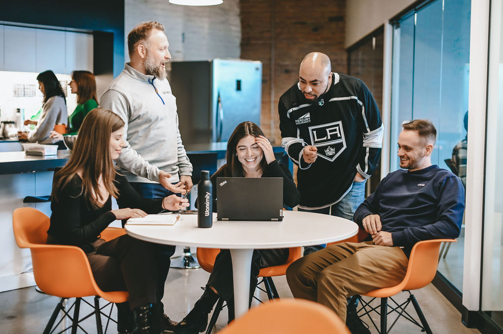 Five colleagues gather around a table, smiling and discussing work on a laptop in a modern office.