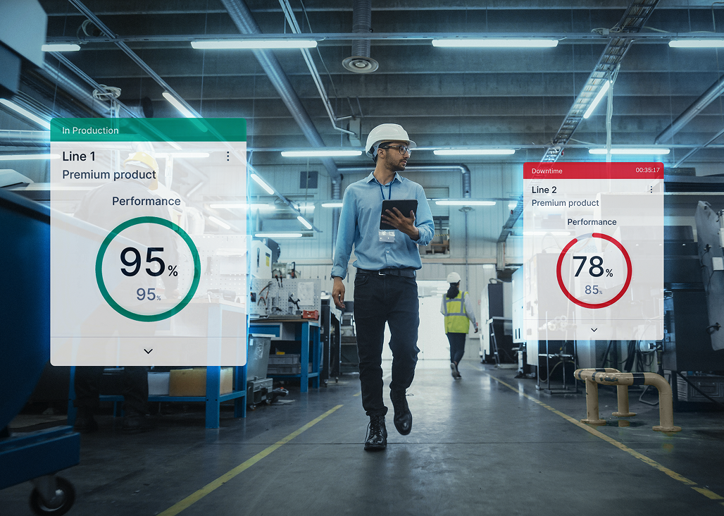 Male factory worker wearing a white hard hat and holding a tablet walking down an aisle in a manufacturing plant with monitoring graphics overlay showing Line 1 at 95% performance in green labeled 'In Production' and Line 2 at 78% performance in red labeled 'Downtime'.