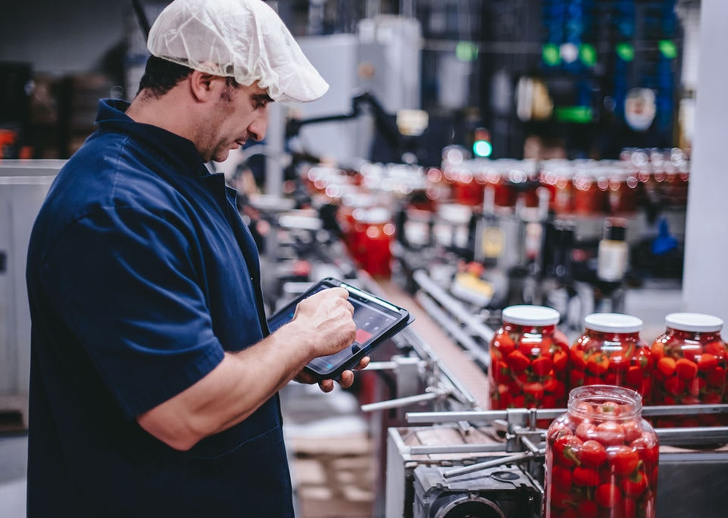 Man in a blue uniform and white hairnet using a tablet to check jars of red peppers on a food production line in a factory setting.
