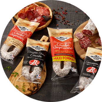 Four packaged French dry sausages labeled 'Saucisson Sec' displayed on wooden boards and a black surface, with some sliced sausage pieces and scattered peppercorns.