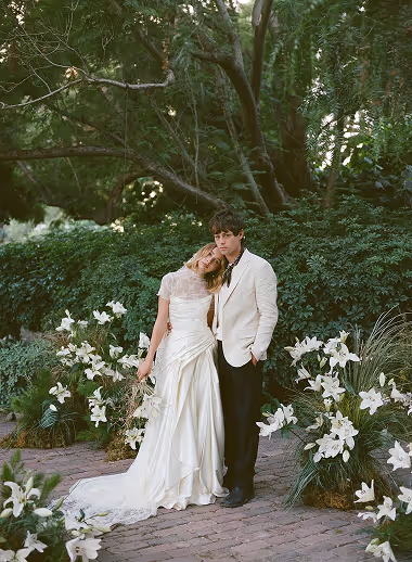 Bride and groom in wedding attire standing amid white lilies and greenery outdoors.
