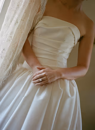 Bride in a strapless satin wedding dress holding her hands with an engagement ring visible.