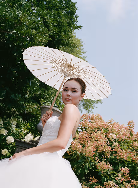 Bride in a white dress holding a white parasol outdoors with green foliage and flowers in the background.