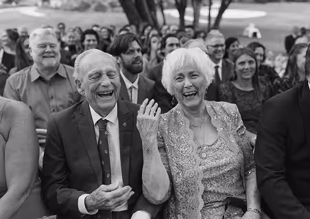 Elderly couple laughing joyfully while seated among guests at an outdoor event.