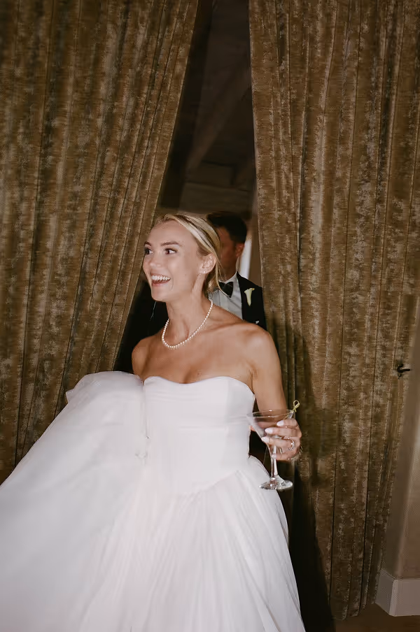 Smiling bride holding her white wedding dress and a cocktail glass, standing in front of brown curtains.