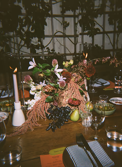 Table centerpiece with mixed flowers, pears, grapes, and lit candles in a cozy dining setting.
