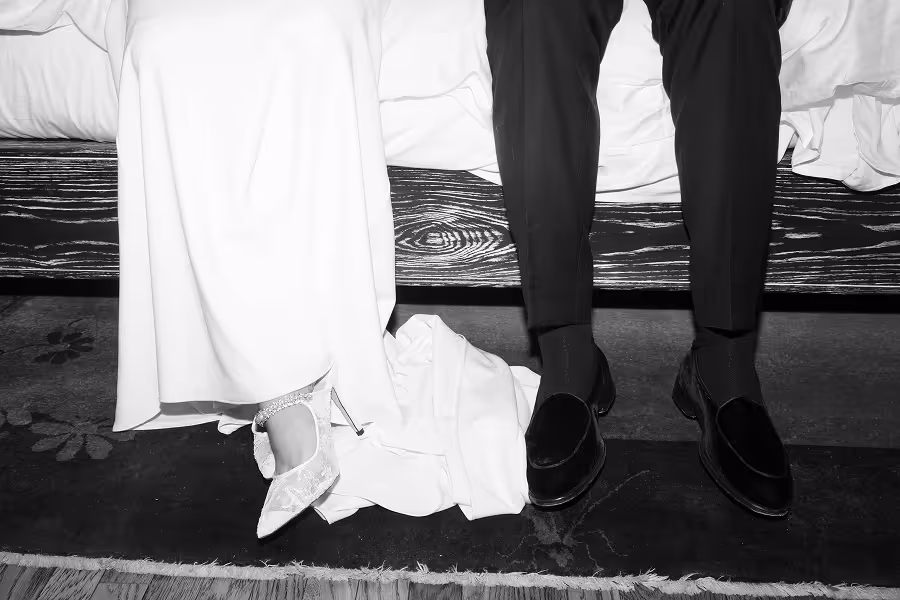 Black and white photo of a couple's lower legs and feet sitting on a bed, woman in heels, man in dress shoes.