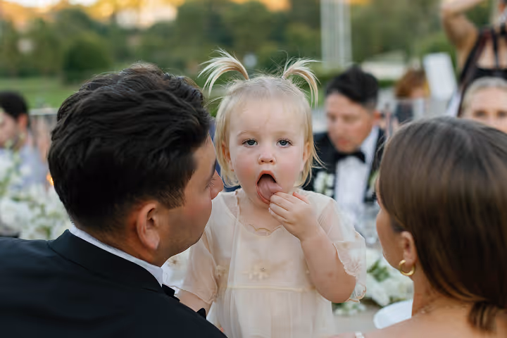 Young girl with pigtails and beige dress sitting between two adults at an outdoor event.