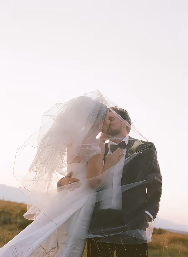Bride and groom kissing outdoors with the bride’s veil flowing around them at sunset.