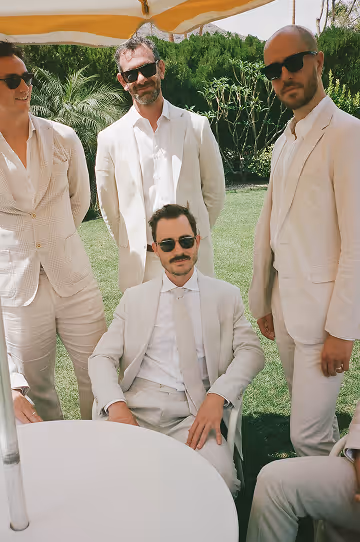 Four men in light-colored suits and sunglasses outdoors near a white table and greenery.