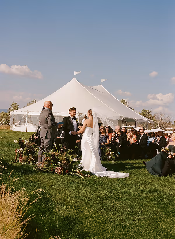 Bride and groom hold hands during outdoor wedding ceremony with white tent and seated guests.