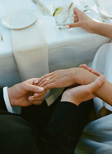 Two people holding hands at a table, one wearing a ring, the other holding a glass with lime and straw.