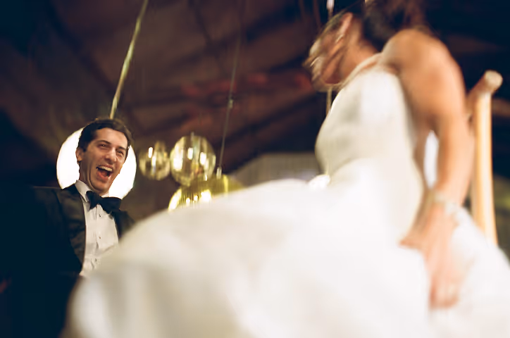 Groom in tuxedo laughing joyfully looking at bride in white wedding dress indoors.