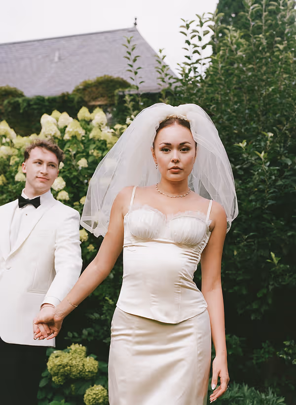 Bride in a white satin dress and veil holding hands with groom in white tuxedo outdoors.