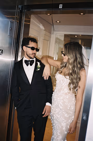 Man in black tuxedo and woman in white floral dress wearing sunglasses inside an elevator.