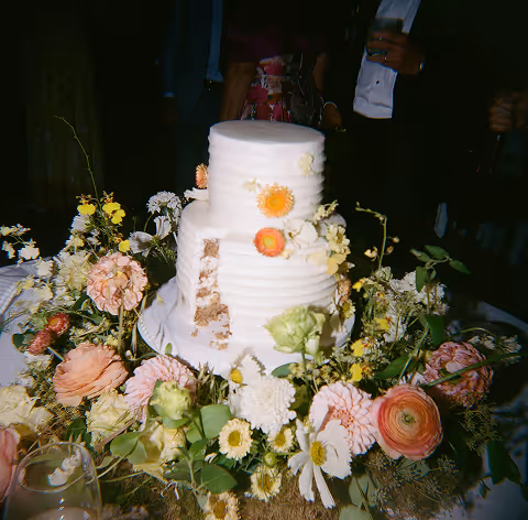 Two-tier white cake with a slice cut, decorated with orange and white flowers and surrounded by assorted flowers.
