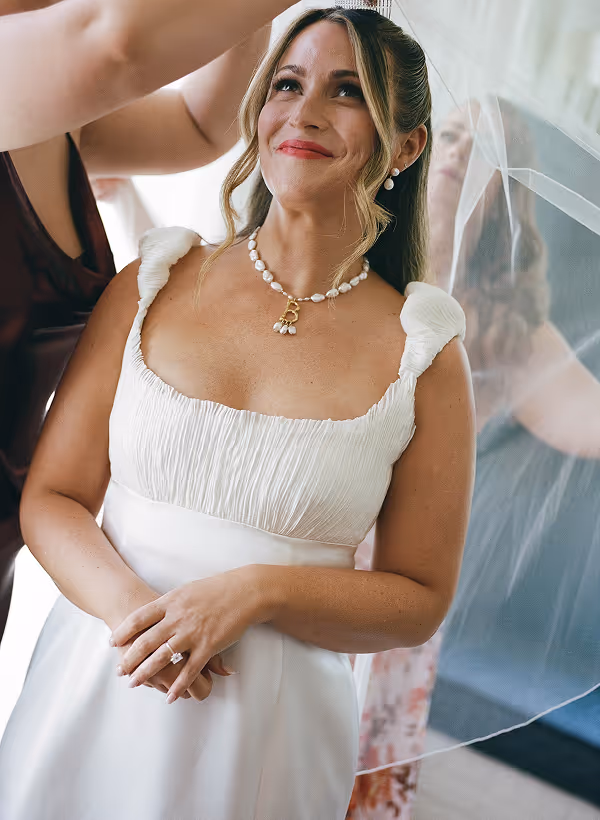 Bride smiling with a pearl necklace and veil being placed on her head.