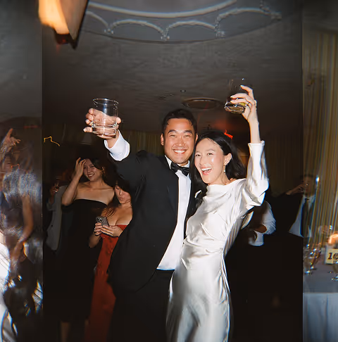 Couple in formal attire smiling and holding drinks up at a party with others in the background.