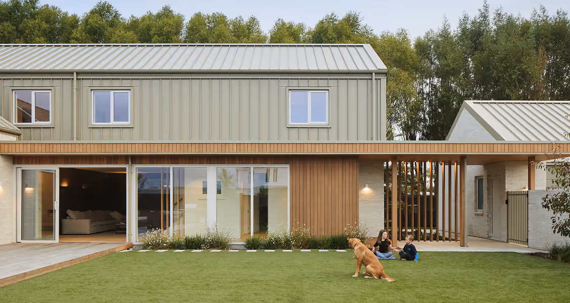 Modern two-story house with large glass doors opening to a lawn where two children sit with a golden retriever.